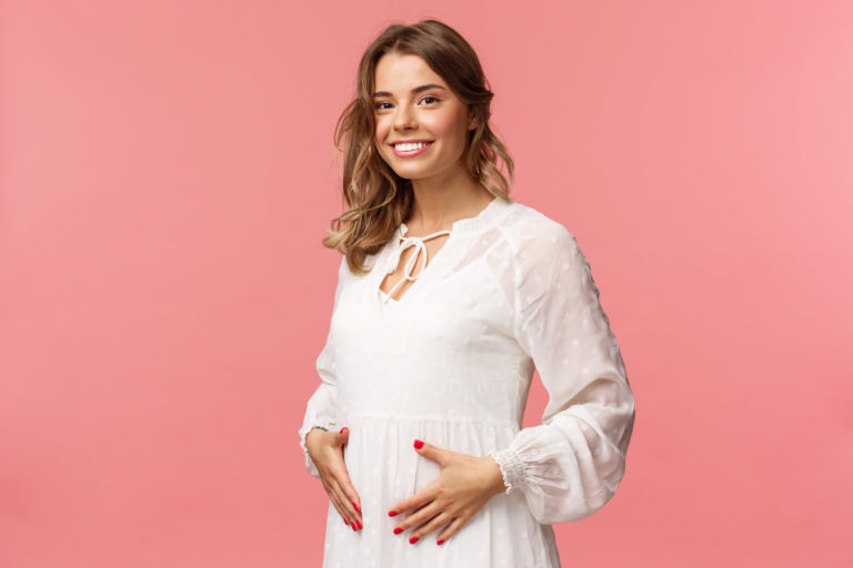 Maternity, women and beauty concept. Tender, cute smiling blond woman in white dress feeling happiness and love, touching belly as expecting child, being pregnant, pink background